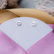 A pair of silver-colored stud earrings with a cool boy emoji design, displayed on a pink cushion.